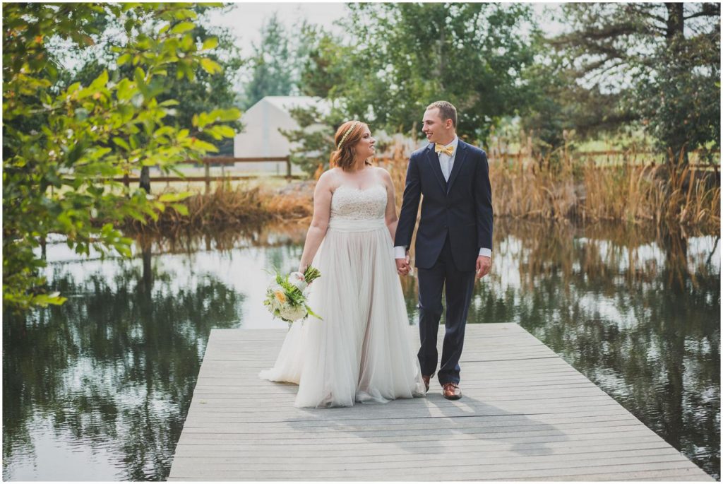 bride and groom walking down pond dock while smiling at each other